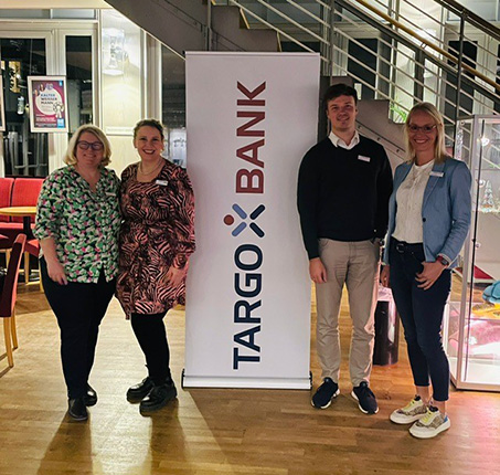 Vier Personen stehen vor einem TARGO BANK Banner in einem Restaurant. Zwei Frauen tragen bunte Oberteile, während zwei Männer formell gekleidet sind.
