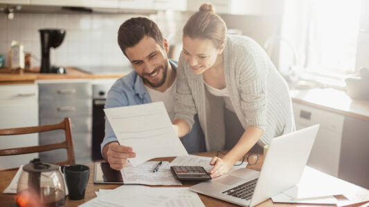 Ein Paar sitzt an einem Küchentisch, umgeben von Dokumenten und einem Laptop. Die Frau zeigt auf ein Blatt Papier, während der Mann es betrachtet. Eine Tasse Kaffee und ein Taschenrechner sind ebenfalls sichtbar.