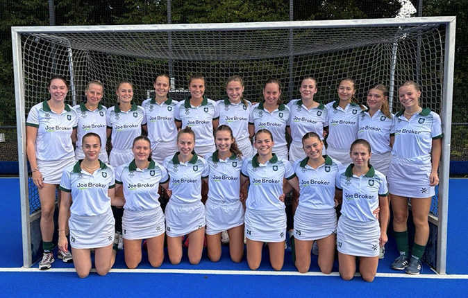 Eine Gruppe von 16 Personen in weißen Sporttrikots mit grünen Kragen steht in einer Reihe vor einem Hockeytor. Die Trikots tragen den Namen "Joe Broker". Der Boden ist blau.