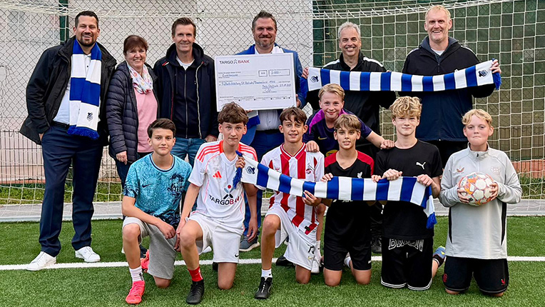 Eine Gruppe von Fußballspielern posiert auf einem Spielfeld. Sie tragen verschiedene Trikots, darunter ein weißes mit roten Streifen. Einige halten Schals mit blauen und weißen Streifen. Im Hintergrund steht eine Gruppe von Erwachsenen, und ein großer Scheck mit der Aufschrift 'TARGOBANK' ist sichtbar.