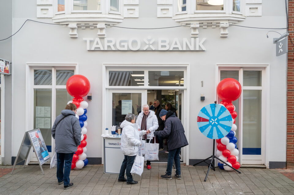 Eine TARGO BANK-Filiale mit einem Eingang, der von roten und blauen Luftballons geschmückt ist. Vor der Bank stehen mehrere Personen, die mit einem Mitarbeiter interagieren. Ein Glücksrad ist sichtbar, das für eine Veranstaltung genutzt wird.