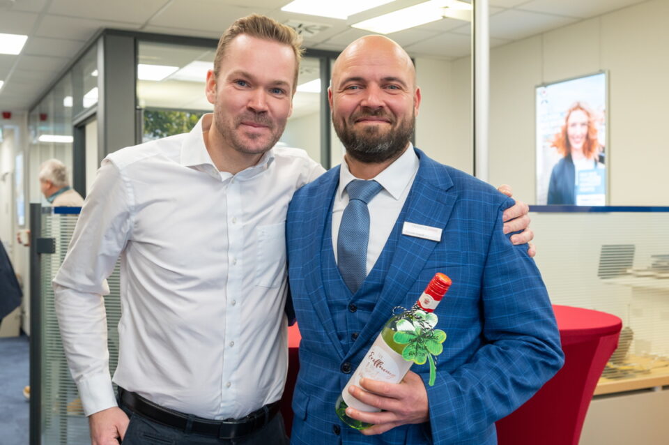 Zwei Männer posieren in einem modernen Büro. Der Mann links trägt ein weißes Hemd, der Mann rechts einen blauen Anzug mit einer Krawatte. Der Mann rechts hält eine Flasche mit rotem Deckel, die mit einem grünen Kleeblatt dekoriert ist. Im Hintergrund sind Möbel und Werbung sichtbar.