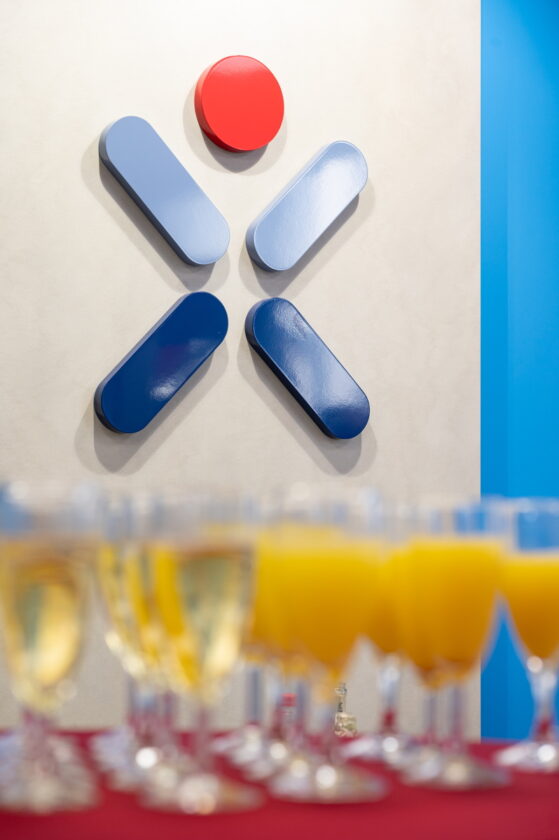 Im Bild sind mehrere Gläser mit Getränken auf einem roten Tisch im Vordergrund zu sehen. Die Gläser enthalten eine gelbe Flüssigkeit sowie einige mit einem klaren Getränk. Im Hintergrund befindet sich eine Wand mit einem modernen, grafischen Design, das einen roten Kreis und mehrere blaue, ovale Formen zeigt, die in einer dynamischen Anordnung angeordnet sind.