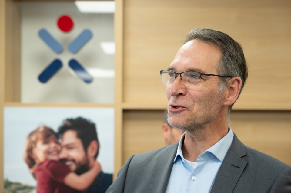 Ein Mann in einem grauen Anzug mit einem hellblauen Hemd steht vor einem Hintergrund aus Holzregalen. Im Hintergrund sind unscharfe Bilder von Personen zu sehen. Links ist ein Firmenlogo mit roten und blauen Elementen sichtbar.