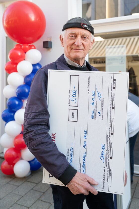 Eine Person hält einen großen Scheck, der auf 5.000 EUR ausgestellt ist. Der Scheck trägt die Aufschrift "Spende" und ist für die Organisation "Kindertafel - Aho Heide" datiert auf den 16.12.2023. Im Hintergrund sind Luftballons in den Farben Rot, Weiß und Blau zu sehen. Die Person trägt einen dunklen Pullover und eine Mütze.