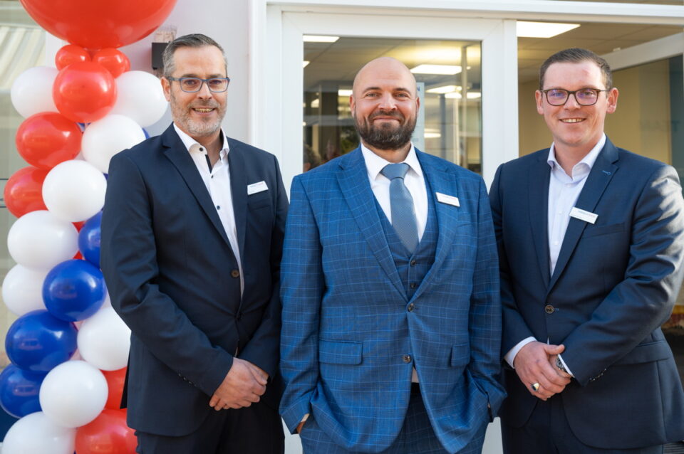 Drei Männer stehen in formeller Kleidung vor einem Gebäude. Der erste Mann trägt einen schwarzen Anzug, der zweite einen blauen karierten Anzug mit Krawatte, und der dritte einen dunklen Anzug. Im Hintergrund sind bunte Luftballons in Rot, Weiß und Blau zu sehen.