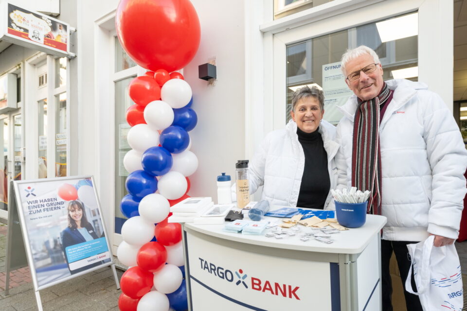 Ein Stand der TARGO BANK draußen, mit zwei Personen in weißen Jacken. Im Hintergrund steht eine Luftballon-Säule in rot, blau und weiß. Auf dem Tisch liegen Broschüren, ein Trinkbehälter und Werbegeschenke. Ein Plakat zeigt den Text: "WIR HABEN EINEN GRUND ZUM FEIERN - Folgen Sie uns und mehr!"