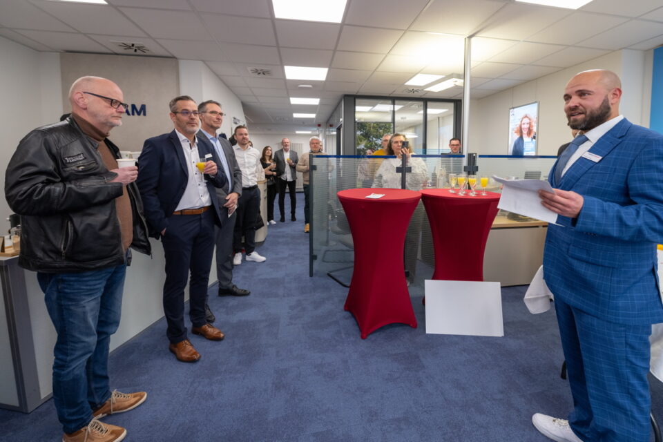 Eine Gruppe von Personen steht in einem modernen Büro. Einige halten Getränke in der Hand. Im Hintergrund sind rot gedeckte Stehtische mit Getränken und ein Bildschirm sichtbar. Die Atmosphäre wirkt geschäftlich und einladend.