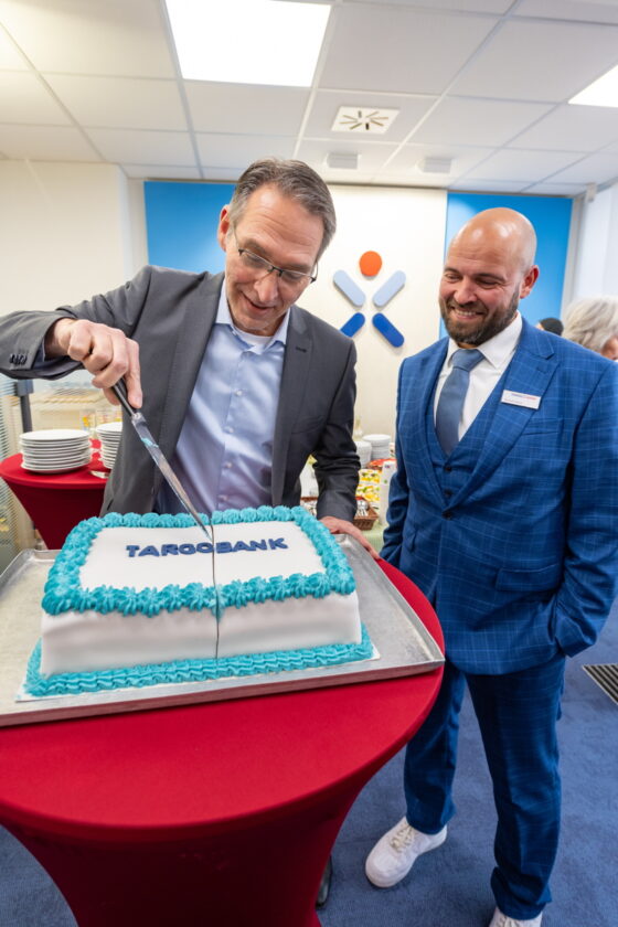 Eine Person schneidet eine große Torte mit dem Schriftzug 'TARGOBANK'. Die Torte hat eine weiße Glasur mit blauen und weißen Dekorationen. Im Hintergrund sind Tische mit Snacks zu sehen. Eine zweite Person steht daneben und trägt einen blauen Anzug.