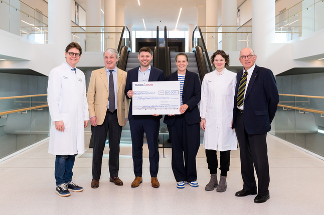 Sechs Personen stehen in einem modernen Innenraum. Sie halten einen großen Scheck mit einer Spendenhöhe. Die Personen tragen verschiedene Kleidung, darunter Laborkittel. Im Hintergrund sind Rolltreppen sichtbar.