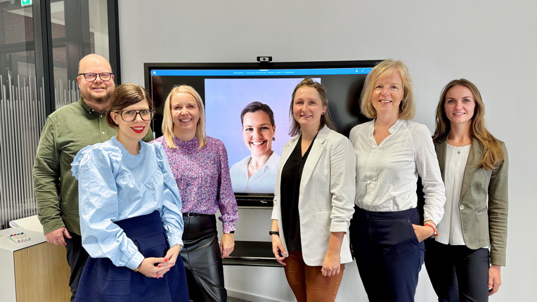 Eine Gruppe von sechs Personen steht vor einem Bildschirm. Die Personen tragen verschiedene Oberbekleidungen, darunter eine blaue Bluse, ein gemustertes Hemd und Blazer. Im Hintergrund ist ein Bildschirm sichtbar, auf dem ein Bild einer weiteren Person zu sehen ist.