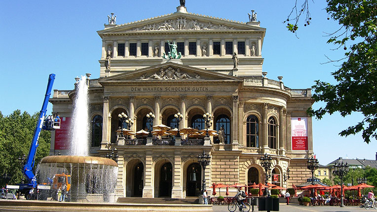 Das Bild zeigt ein beeindruckendes historisches Theatergebäude mit einem großen Brunnen im Vordergrund. Auf der Vorderseite des Gebäudes sind große Fenster und Säulen zu sehen, mit Terrassen, auf denen Gäste sitzen. Ein Kran und ein orangefarbenes Fahrzeug sind sichtbar, die an der Renovierung oder Wartung des Gebäudes beteiligt sind. Im Hintergrund sind Bäume und weitere Terrassen mit roten Sonnenschirmen zu sehen.