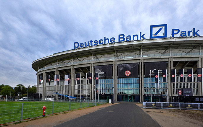 Das Bild zeigt das Deutsche Bank Park Stadion. Im Vordergrund ist der Eingang des Stadions mit großen Fenstern und einem breiten Gehweg sichtbar. Über dem Stadion steht der Name "Deutsche Bank Park" in großen Buchstaben. Auf der linken Seite ist ein grünes Spielfeld zu sehen, und auf der rechten Seite stehen Werbebanner. Der Himmel ist bewölkt.