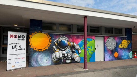 Eine Wand mit einem bunten Graffiti zeigt einen Astronauten, die Erde, die Sonne, den Mond und mehrere Planeten. Ein Banner mit der Aufschrift "NO MOBBING - Wir sagen NEIN zu Mobbing" steht daneben.