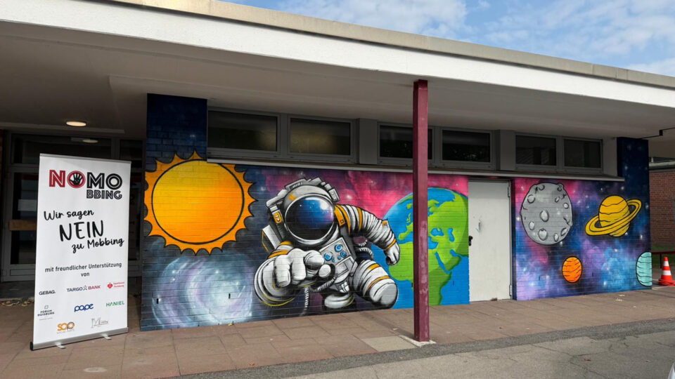 Eine Wand mit einem bunten Graffiti zeigt einen Astronauten, die Erde, die Sonne, den Mond und mehrere Planeten. Ein Banner mit der Aufschrift "NO MOBBING - Wir sagen NEIN zu Mobbing" steht daneben.