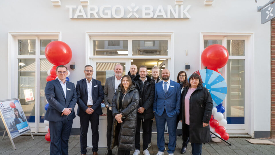 Eine Gruppe von Menschen steht vor einer TARGOBANK-Filiale. Sie tragen formelle Kleidung und lächeln. Im Hintergrund sind rote und blaue Luftballons sowie ein Glücksrad sichtbar. Ein Plakat steht links neben der Tür.