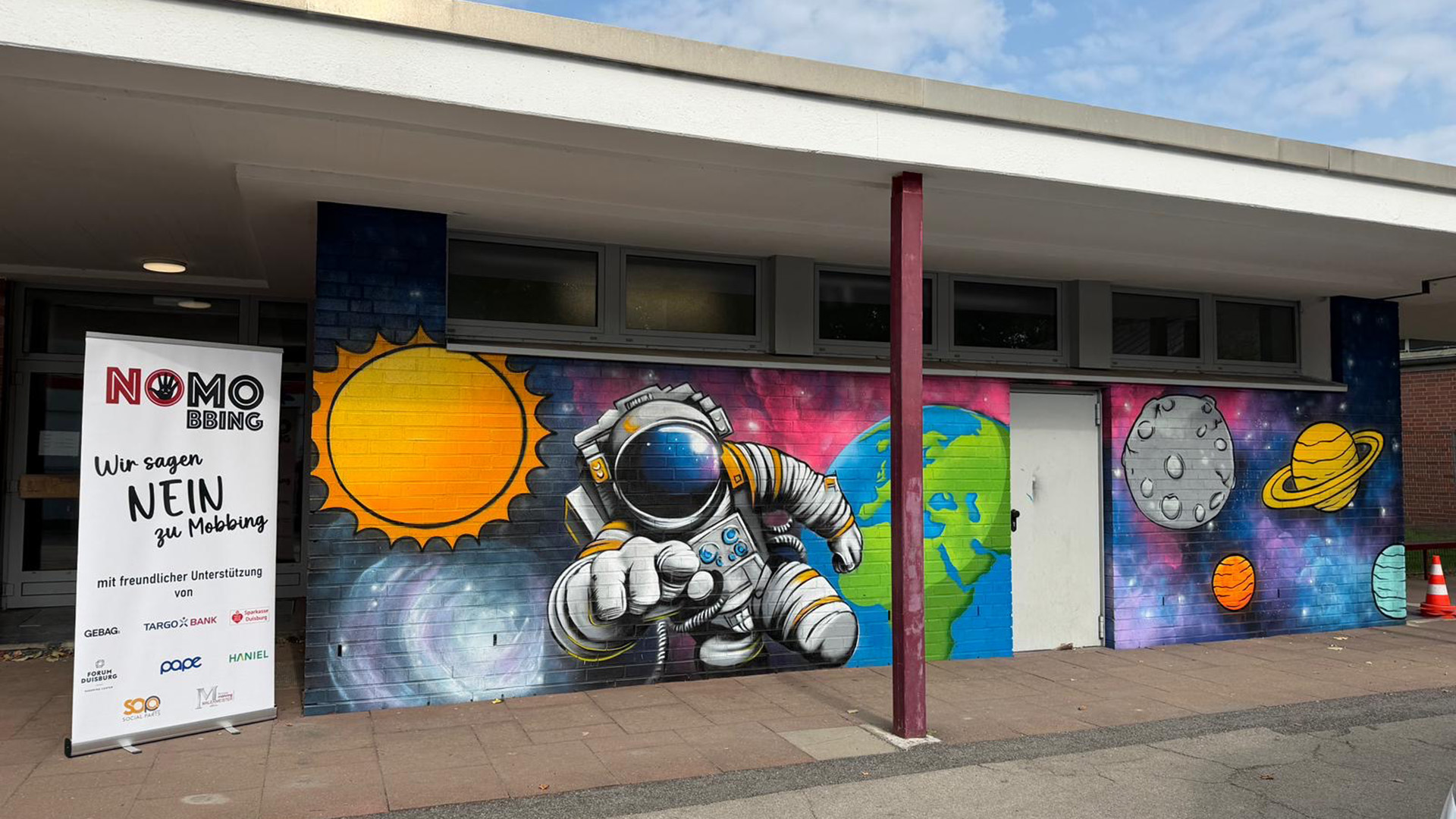 Eine Wand mit einem bunten Graffiti zeigt einen Astronauten, die Erde, die Sonne, den Mond und mehrere Planeten. Ein Banner mit der Aufschrift 