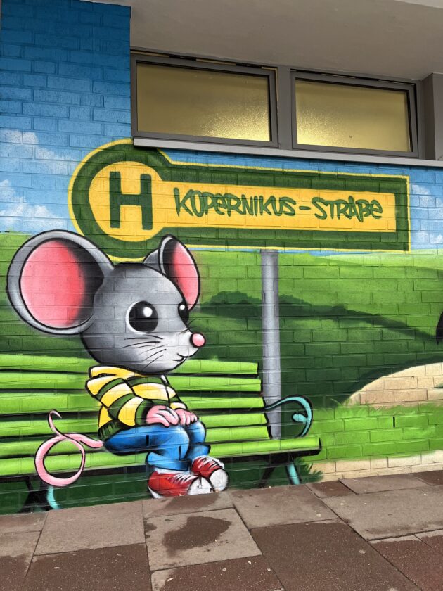 Ein Graffiti auf einer Wand zeigt eine cartoonartige Maus, die auf einer grünen Bank sitzt. Die Maus trägt ein gestreiftes gelbes Hoodie und blaue Jeans. Über der Maus steht ein Schild mit dem Text "KOPERNIKUS-STRAßE". Die Hintergrundfarbe ist hellblau und grün.