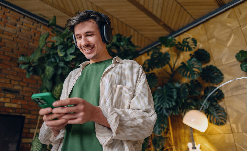 Eine Person steht in einem Raum mit Pflanzen im Hintergrund, trägt Kopfhörer und hält ein Smartphone in der Hand.