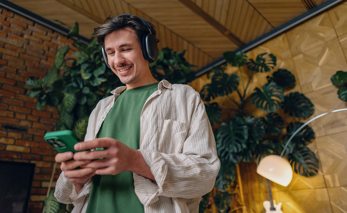 Eine Person steht in einem Raum mit Pflanzen im Hintergrund, trägt Kopfhörer und hält ein Smartphone in der Hand.
