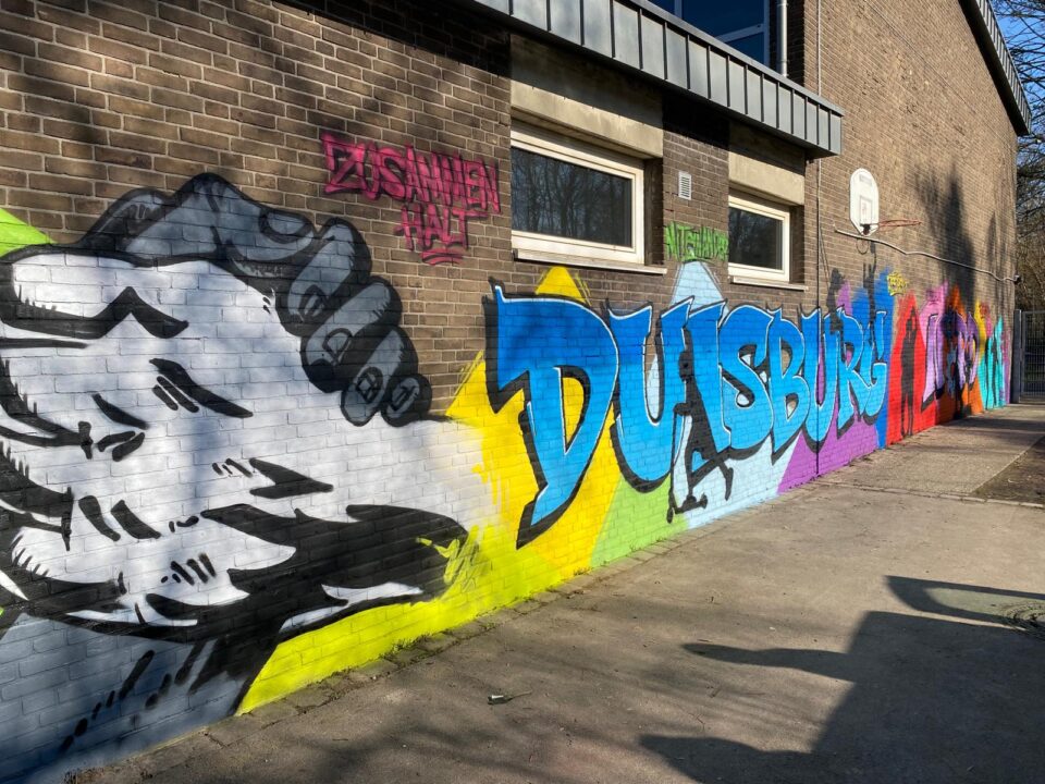 Wandgemälde an einem Gebäude in Duisburg zeigt eine große Hand, die eine andere greift, mit dem Text "ZUSAMMEN HALT" in Pink und bunten Graffiti in blau, grün, gelb und rot.