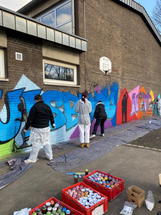 Drei Personen in Schutzanzügen malen ein Wandgemälde auf einer Ziegelwand. Eine Person trägt eine schwarze Jacke, eine andere eine graue. Es gibt zahlreiche Spraydosen in Kisten auf dem Boden.
