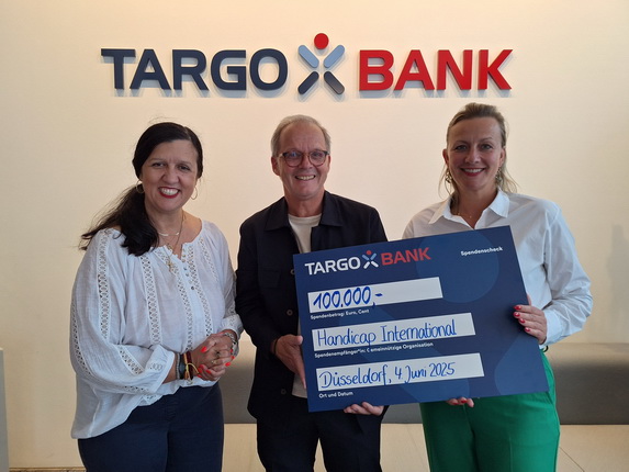 Drei Personen halten einen großen Scheck mit der Aufschrift 100.000 Euro für Handicap International vor dem TARGOBANK-Logo in Düsseldorf.