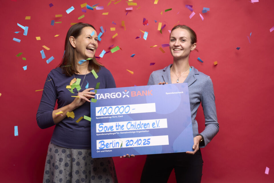 Zwei Personen halten einen großen Scheck mit der Aufschrift "100.000€" für "Save the Children e.V." vor einem roten Hintergrund, umgeben von Konfetti.