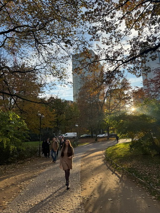 Eine Frau mit langen Haaren in einem beigen Mantel geht auf einem mit Steinen gepflasterten Weg in einem Park. Im Hintergrund sind Bäume mit herbstlichen Blättern und moderne Gebäude sichtbar. Weitere Personen sind am Weg entlang zu sehen.