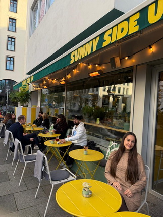 Ein Café mit der Aufschrift "SUNNY SIDE UP" im Freien. Tische und Stühle in Gelb und Grau sind auf dem Gehweg platziert. Gäste sitzen und genießen ihre Getränke und Speisen. Es gibt Pflanzen und Lichter, die eine einladende Atmosphäre schaffen.