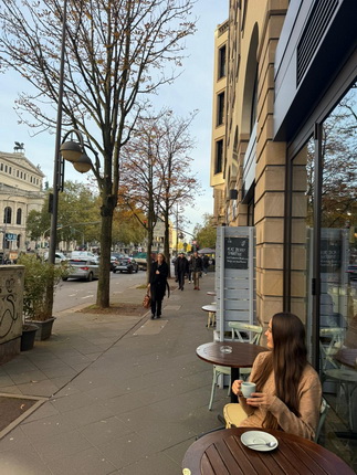 Eine belebte Straße mit Bäumen und Cafés. Eine Frau sitzt an einem Tisch im Freien und hält eine Tasse. Im Hintergrund laufen Passanten.