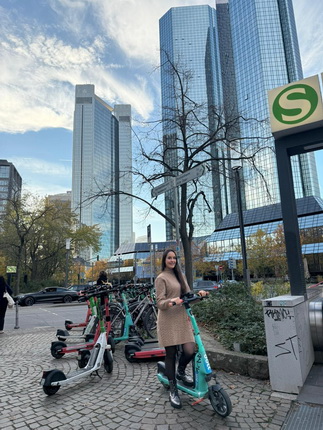 Eine Frau steht auf einem E-Scooter in einer urbanen Umgebung mit modernen Hochhäusern im Hintergrund. Die Frau trägt ein beigefarbenes Kleid und schwarze Stiefel. Im Vordergrund sind mehrere E-Scooter geparkt. Der Himmel ist teilweise bewölkt und es ist Herbst, was an den Bäumen und dem Laub zu erkennen ist. Ein Schild mit einem 'S' ist ebenfalls sichtbar.