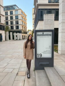 Eine Frau steht lächelnd vor einem modernen Gebäude mit dem Schriftzug 'mainBuilding' und der Nummer 19. Sie trägt ein braunes, gestricktes Kleid und schwarze Stiefel. Der Platz ist gepflastert und umgeben von weiteren modernen Gebäuden.