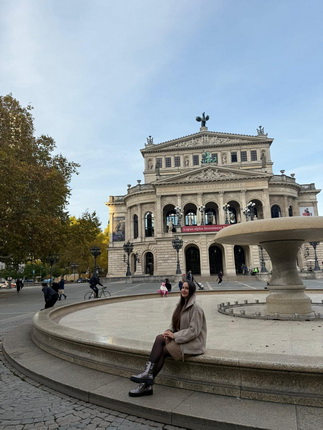 Eine Frau sitzt auf dem Rand eines Brunnens vor einem historischen Gebäude mit Säulen und einer Statue auf dem Dach. Im Hintergrund sind Bäume und Passanten zu sehen. Die Frau trägt einen beigen Mantel und schwarze Stiefel, während der Himmel leicht bewölkt ist.