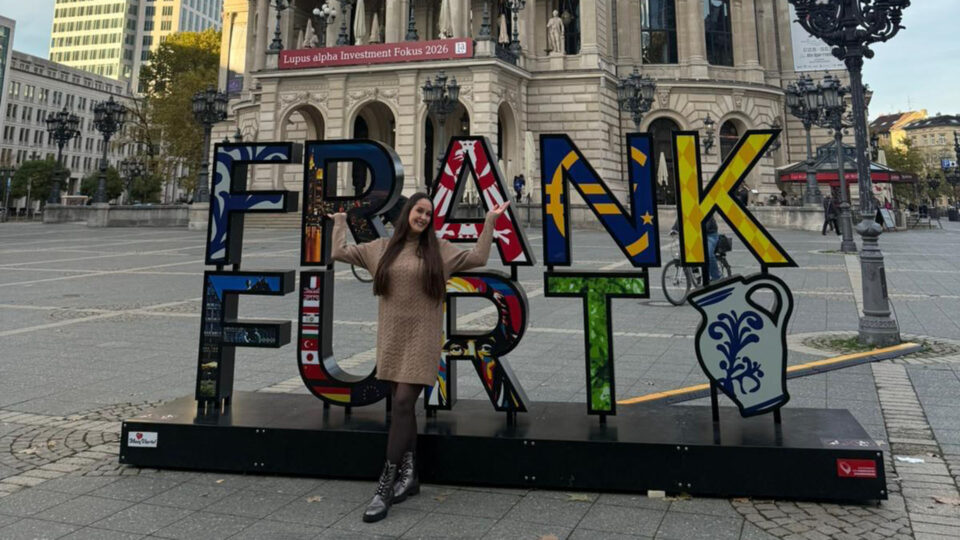 Eine Person steht vor einem bunten Schriftzug "FRANKFURT" in einer städtischen Umgebung. Der Schriftzug enthält verschiedene Designs und Flaggen. Im Hintergrund ist ein historisches Gebäude mit Statuen und Laternen sichtbar. Es sind weitere Personen und Fahrräder in der Umgebung zu sehen.