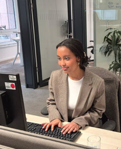 Eine Person sitzt an einem Schreibtisch und arbeitet an einem Computer. Sie trägt einen blauen Blazer über einem weißen Oberteil. Der Schreibtisch ist ordentlich, mit einer Tastatur und einer Glaswasserflasche. Im Hintergrund sind Büropflanzen und ein heller Raum mit Fenstern sichtbar.
