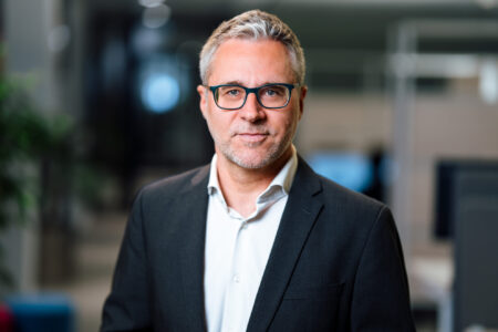 Ein Mann in einem Anzug steht in einem modernen Büro. Im Hintergrund sind verschwommene Büroelemente und Pflanzen sichtbar.