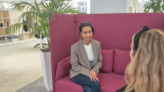 Eine Person sitzt auf einem lila Sofa in einem modernen Büro, umgeben von Pflanzen. Im Hintergrund sind Büromöbel und ein Schreibtisch sichtbar.