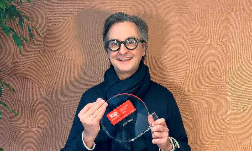 Eine Person hält eine klare, runde Platte in der linken Hand und eine rote Karte in der rechten Hand. Die Karte hat das Logo 'tap' und einige Informationen darauf. Die Person trägt einen dunklen Pullover und einen Schal. Im Hintergrund ist eine strukturierte Wand in warmen, erdigen Tönen zu sehen.