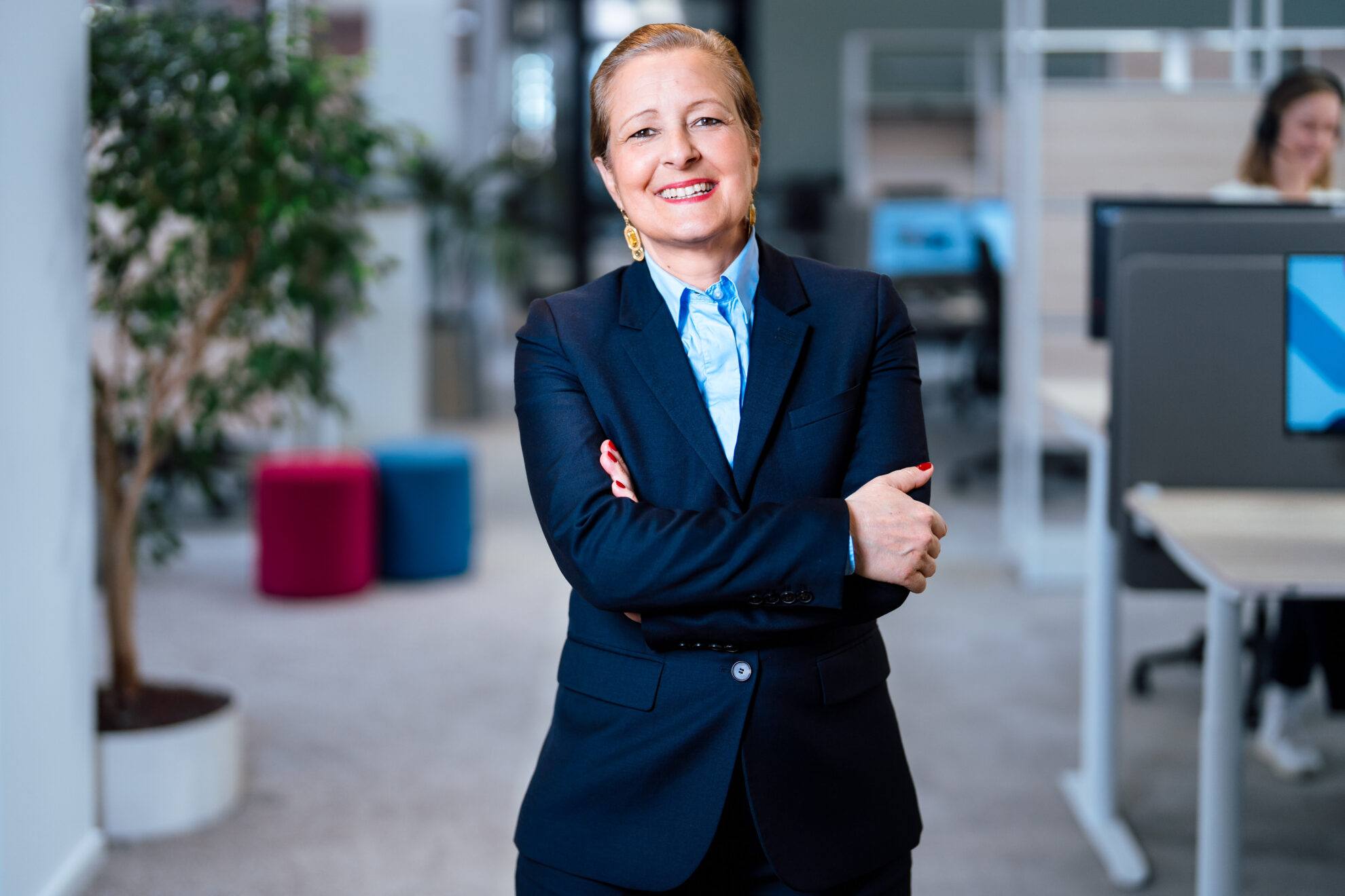 Eine Person in einem blauen Anzug mit verschränkten Armen steht in einem modernen Büro. Im Hintergrund sind Pflanzen und Büroeinrichtungen sichtbar, darunter farbige Hocker und Schreibtische mit Computern.