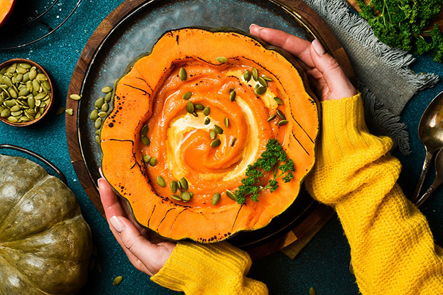 Eine Person mit einer gelben Strickjacke hält eine Schüssel aus Kürbis, gefüllt mit einer Kürbissuppe. Die Suppe hat eine cremige, spiralförmige Garnitur und ist mit Kürbiskernen und frischer Petersilie dekoriert. Auf einem blauen Tisch liegen neben der Schüssel ein paar Kürbiskerne und ein grauer Kürbis.