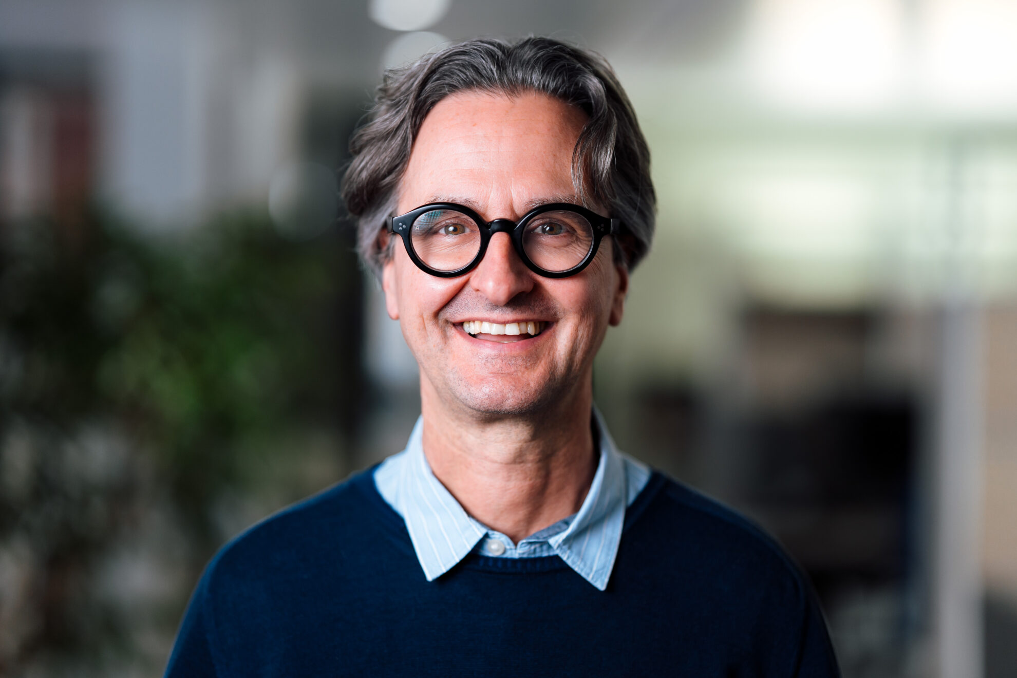 Ein Mann mit grauen Haaren steht in einem modernen Büro. Er trägt einen blauen Pullover über einem hellblauen Hemd. Der Hintergrund ist unscharf und zeigt Anzeichen eines geschäftlichen Umfelds.