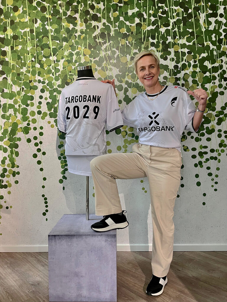 Eine Person steht neben einem Mannequin, das ein weißes Trikot mit der Aufschrift 'TARGOBANK 2029' trägt. Die Person trägt ebenfalls ein Trikot mit dem gleichen Design und hat eine Hand in die Hüfte gestützt. Der Hintergrund zeigt eine grüne, blattartige Wandgestaltung. Die Person trägt beige Hosen und schwarze Schuhe.