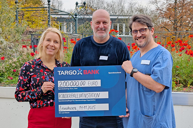 Drei Personen stehen im Freien und halten ein großes Scheck über 200.000 Euro für die Kinderpalliativstation. Die TARGOBANK ist als Sponsor aufgeführt. Im Hintergrund sind bunte Blumen zu sehen. Das Datum ist der 14.11.2025, und der Ort ist Leverkusen.