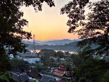 Ein malerischer Blick auf eine Landschaft bei Sonnenaufgang. Im Vordergrund sind Dächer von Häusern sichtbar, umgeben von Bäumen. Im Hintergrund erstrecken sich sanfte Hügel und Berge, während die Sonne gerade aufgeht und den Himmel in sanften Farben erstrahlen lässt. Ein Mobilfunkmast ist zu sehen, der in die Landschaft hineinragt.