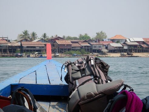 Das Bild zeigt die Vorderansicht eines Bootes, das auf einem Gewässer fährt. Im Vordergrund sind Rucksäcke zu sehen, die auf dem Boot liegen. Im Hintergrund erkennt man ein Dorf mit mehreren Häusern, die ein schräges Dach haben. Palmen sind ebenfalls sichtbar, und der Himmel ist klar und blau.