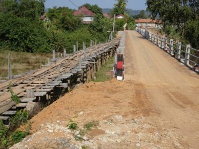 Ein schmaler, holzgeplankter Weg führt über einen Fluss. Auf der linken Seite ist eine einfache Holzbrücke zu sehen, die einige unregelmäßig angeordnete Holzplanken aufweist. Rechts daneben verläuft eine befestigte Straße, die gut gepflegt aussieht. Im Hintergrund sind grüne Bäume und einige Häuser sichtbar. Der Himmel ist klar und sonnig, was auf einen schönen Tag hinweist.