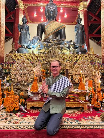 Eine Person kniet in einem Tempel vor einer großen Buddha-Statue. Die Statue ist von mehreren weiteren Statuen umgeben, und der Raum ist reich verziert. Vor der Person stehen Opfergaben, darunter orangefarbene Blumen und Kerzen, auf einem prächtigen Teppich. Die Person trägt ein grünes Hemd und hat einen traditionellen Schal über der Schulter.