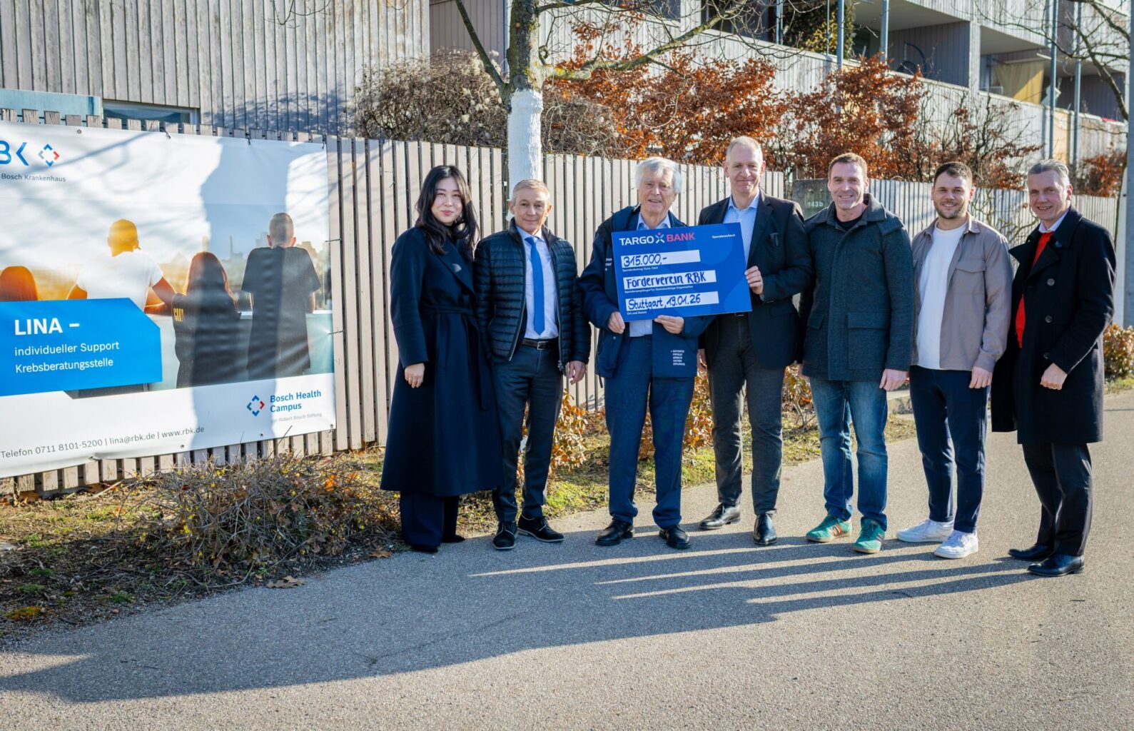 Eine Gruppe von Personen steht vor einem Banner, das Informationen über die Krebsberatungsstelle LINA enthält. Sie halten einen großen Scheck von 315.000 Euro für den Förderverein RBK. Der Hintergrund zeigt ein modernes Gebäude und Bäume, die im Winter keine Blätter haben.