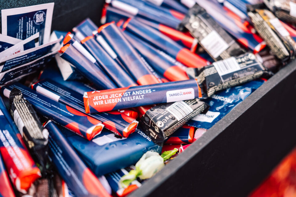 Ein Haufen von bunten Verpackungen, darunter einige mit dem Schriftzug "JEDER JECK IST ANDERS – WIR LIEBEN VIELFALT" und dem Logo der TARGOBANK. Die Verpackungen sind größtenteils in Blau und Rot gehalten und liegen in einer schwarzen Box.